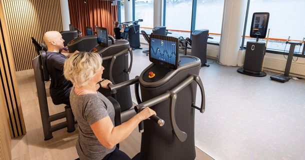 Innenansicht eines modernen Fitnessstudios in Finnland. Im Vordergrund trainiert eine Frau an einem dunklen EGYM-Kraftgerät mit großem Display, das eine Wellenkurve zur Bewegungsführung zeigt. Im Hintergrund sind weitere vernetzte Trainingsgeräte und eine Fensterfront zu sehen.