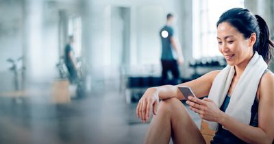 Frau mit Handtuch um den Schultern hält Lächelnd ein Handy in einem Gym in der Hand