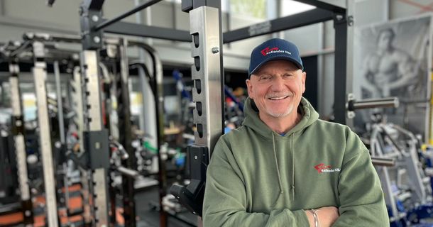 Ein lächelnder Mann in Realleader-USA-Kleidung lehnt vor einem Power Rack im Fitnessstudio.
