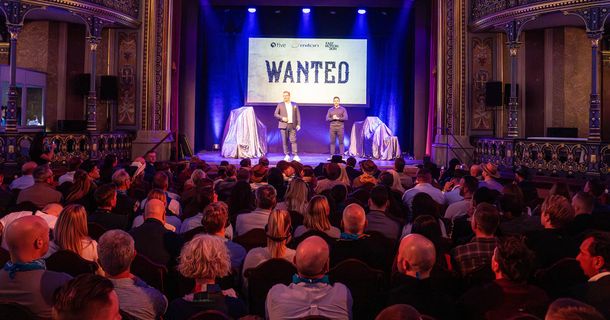 Publikum blickt auf die Bühne, auf der Wolf Harwath und Bernhard-Stefan Müller stehen, auf der Leinwand dahinter steht "WANTED"