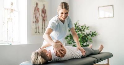 Eine Physiotherapeutin in hellblauer Arbeitskleidung behandelt eine Patientin, die auf einer Behandlungsliege liegt. Die Therapeutin lächelt und mobilisiert mit ihren Händen die Schulter- und Rückenpartie der Frau. Im hellen Behandlungsraum sind im Hintergrund ein anatomisches Poster und ein Skelettmodell zu sehen
