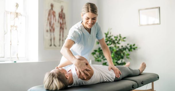 Eine Physiotherapeutin in hellblauer Arbeitskleidung behandelt eine Patientin, die auf einer Behandlungsliege liegt. Die Therapeutin lächelt und mobilisiert mit ihren Händen die Schulter- und Rückenpartie der Frau. Im hellen Behandlungsraum sind im Hintergrund ein anatomisches Poster und ein Skelettmodell zu sehen