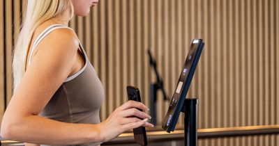Eine blonde Frau in grauer Sportkleidung steht vor einer Holzlamellenwand und hält ihr Smartphone an das Terminal des biometrischen Facetronic IDA-Zutrittslesers.