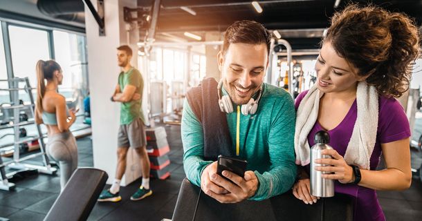 Zwei Frauen und zwei Männer befinden sie in einem Fitnessstudio. Der Mann im Vordergrund zeigt einer Frau etwas auf seinem Smartphone.