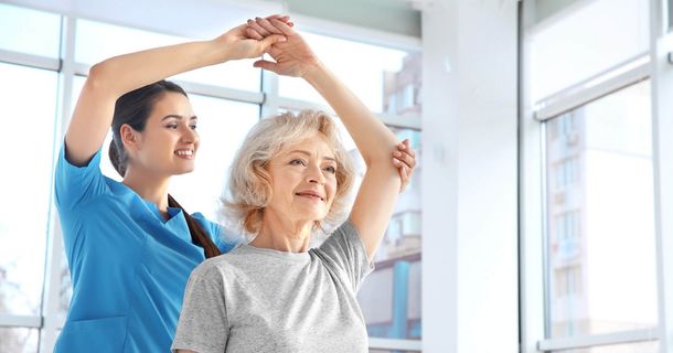 Ältre Dame bewegt ihren Arm über den Kopf mit Hilfe einer Therapeutin