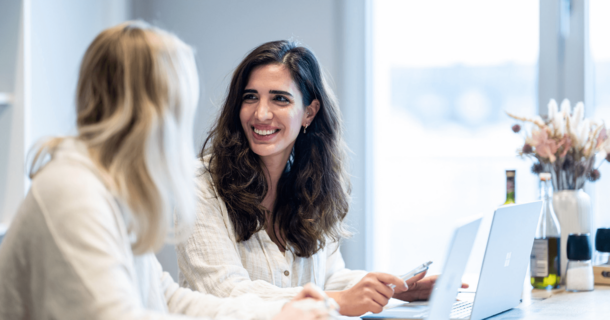Zwei Frauen sitzen vor ihren Laptops und lächeln sie an.