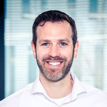 Aggregatoren im Check: Wellhub & EGYM Wellpass beziehen Stellung -1 Porträt eines lächelnden Mannes mit kurzen, dunklen Haaren und Bart, der ein weißes Hemd trägt. Er blickt direkt in die Kamera vor einem hellen, leicht unscharfen Hintergrund, der an ein modernes Bürogebäude erinnert.