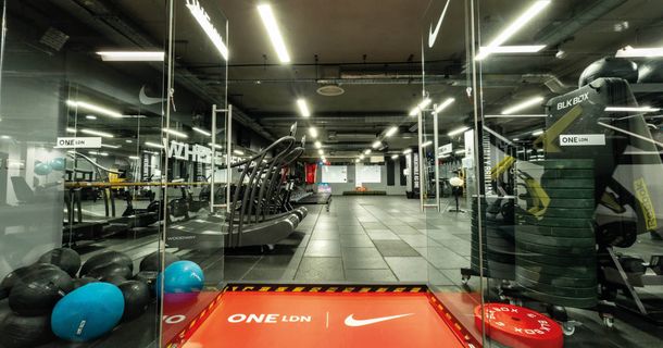 Blick durch eine geöffnete Glastür in ein modernes Fitnessstudio mit roter ONE LDN x Nike-Fußmatte. Im Vordergrund liegen Medizinbälle, rechts stehen Hantelscheibenstapel und Racks von BLK BOX, im Hintergrund befinden sich gebogene Laufbänder unter hellem Deckenlicht.