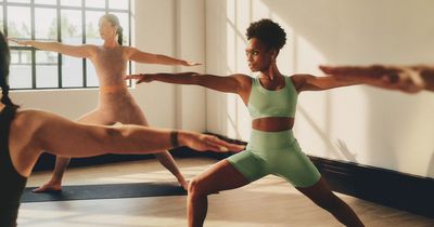 Mehrere Frauen absolvieren barfuß auf Yogamatten einen Yoga-Kurs. Im Fokus steht eine Frau in hellgrünem Sport-Set in der Krieger-Pose (Virabhadrasana), während im Hintergrund vor einem sprossenlosen Fenster weitere Kursteilnehmerinnen dieselbe Bewegung ausführen.