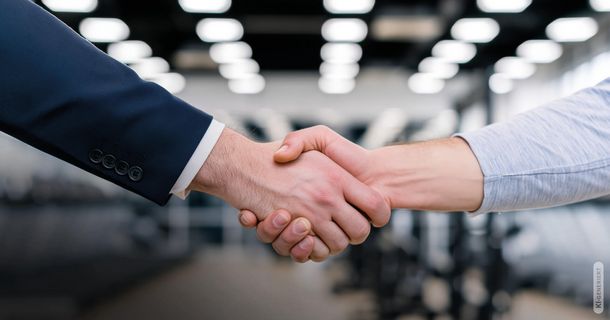 Nahaufnahme von zwei Händen beim Händedruck: Links ein Ärmel eines dunklen Anzugs mit weißer Manschette, rechts ein hellgraues Oberteil. Der Hintergrund zeigt verschwommen die beleuchtete Trainingsfläche eines Fitnessstudios.