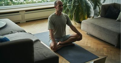 Mann sitzt im Wohnzimmer im Schneidersitz vor dem Laptop und meditiert