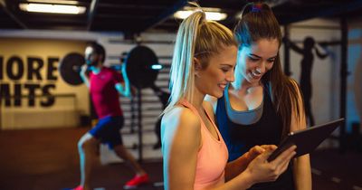 In einem Fitnessstudio steht eine Mitarbeiterin mit einem Tablett in der Hand neben einer Trainierenden.