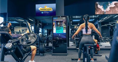 Eine Frau auf einem Fahrrad-Ergometer und ein Mann an einem Kraftgerät trainieren im Fitnessstudio. Im Zentrum steht eine hochformatige, digitale airtango-Stele, im Hintergrund hängt ein TV-Bildschirm mit airtango-Entertainment.