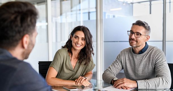 Drei Personen sitzen an einem gläsernen Besprechungstisch in einem modernen Büro. Im Zentrum sitzen eine lächelnde Frau mit dunklem, langem Haar (grünes Oberteil) und ein Mann mit Brille (grauer Pullover), die einem dritten, mit dem Rücken zur Kamera sitzenden Gesprächspartner zuhören oder mit ihm verhandeln. Auf dem Tisch liegen Dokumente und ein Smartphone.