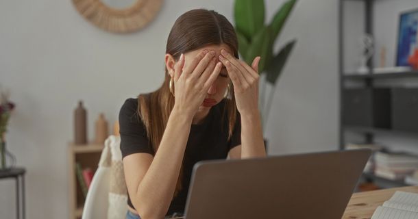 Eine junge Frau sitzt erschöpft vor ihrem Laptop an einem Schreibtisch und hält sich mit beiden Händen die Stirn. Sie wirkt gestresst oder konzentriert. Im Hintergrund ist eine helle, modern eingerichtete Wohnung mit Pflanzen und Regalen zu sehen.