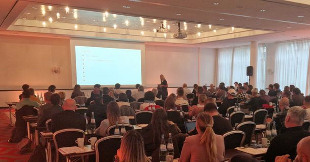 Volles Haus beim Finale der myline Livetour - Header Eine gut besuchte Konferenz oder Schulung in einem großen Saal. Zahlreiche Teilnehmer sitzen in Reihen an Tischen mit Wasserflaschen und Kaffeebechern und blicken auf eine Leinwand mit einer Präsentation. Eine Person steht vorne am Rednerpult. Der Raum hat eine rote Teppichboden und hohe Fenster mit weißen Vorhängen. Die Szene zeigt das Finale der myline-Livetour.