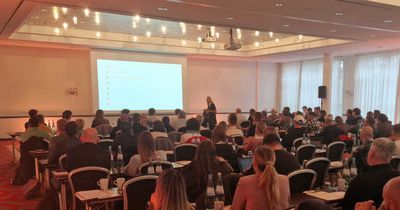 Eine gut besuchte Konferenz oder Schulung in einem großen Saal. Zahlreiche Teilnehmer sitzen in Reihen an Tischen mit Wasserflaschen und Kaffeebechern und blicken auf eine Leinwand mit einer Präsentation. Eine Person steht vorne am Rednerpult. Der Raum hat eine rote Teppichboden und hohe Fenster mit weißen Vorhängen. Die Szene zeigt das Finale der myline-Livetour.