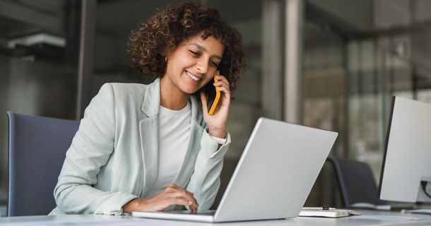 Eine Frau mit dunklen Locken und hellgrauem Blazer sitzt an einem hellen Schreibtisch. Sie blickt lächelnd auf ihren geöffneten Laptop, während sie mit einem Smartphone am Ohr telefoniert.