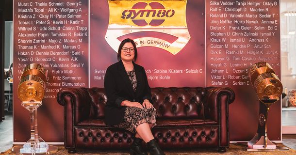 Samantha Timms sitzt auf einem Sofa, im Hintergrund ist eine weinrote Wand mit goldenem gym80-Logo