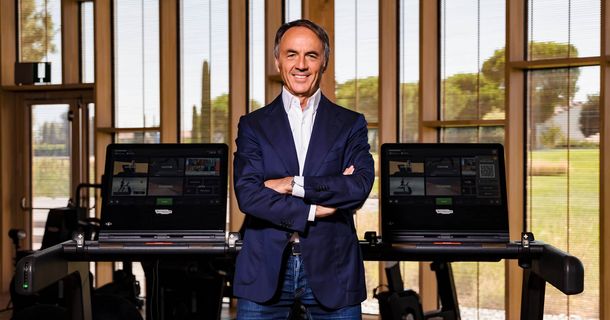 Der Technogym-Gründer und -Präsident Nerio Alessandri steht lächelnd mit verschränkten Armen zwischen zwei seiner hochmodernen, vernetzten Fitnessgeräte. Er trägt einen marineblauen Anzug über einem weißen Hemd und Jeans. Im hellen Hintergrund ist eine große Fensterfront zu sehen, die auf eine begrünte Außenanlage blickt, was eine Wellness-Campus-Atmosphäre vermittelt.