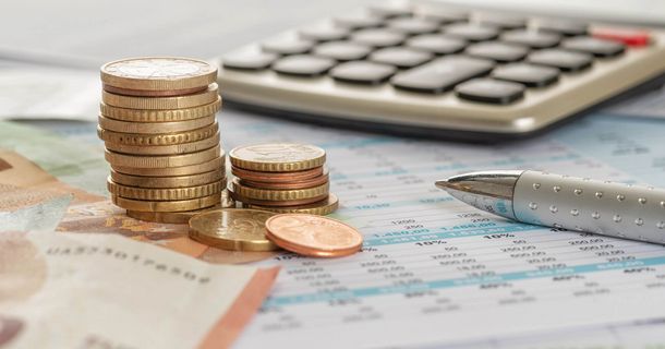 Nahaufnahme von zusammengestapelten Euro-Münzen und Geldscheinen, die neben einem silbernen Kugelschreiber auf einem Finanzdokument mit Tabellen liegen. Im unscharfen Hintergrund ist ein Tischrechner zu sehen.