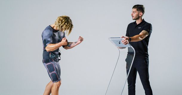 Ein EMS-Trainer hält seine Hände auf einem EMS Tableau und gegenüber ist ein Trainierender der seine Fäuste anspant und einen DrySuit von XBody trägt.