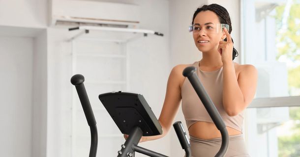 Eine junge Frau in beige-farbener Sportbekleidung steht auf einem Crosstrainer und berührt mit der Hand den Bügel einer futuristischen, transparenten Smart Glasses, um diese zu bedienen.