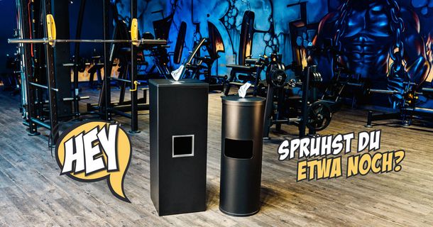 Zwei schwarze Hygiene-Spendersäulen für Desinfektionstücher stehen auf dem Holzfußboden eines Fitnessstudios.