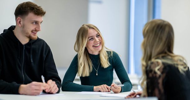 Ein junger Mann und zwei junge Frauen sitzen am Tisch, unterhalten sich und lachen dabei 