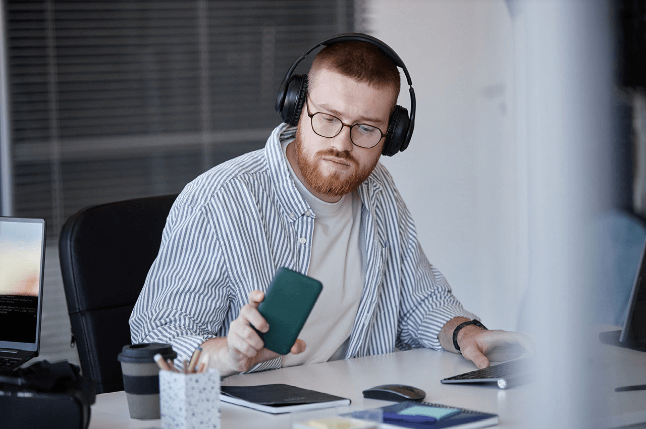 Ein Mitarbeiter sitzt an seinem Schreibtisch und hält ein Smartphone in der Hand auf das er blickt.