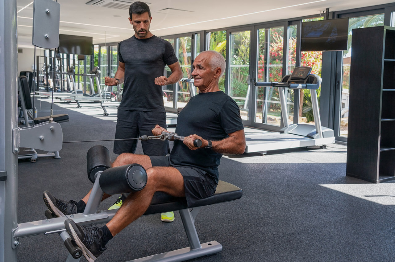 Ein &auml;lterer Mann trainiert an einer Rudermaschine im Fitnessstudio, w&auml;hrend ein Trainer daneben steht und die Ausf&uuml;hrung der &Uuml;bung aufmerksam begleitet.