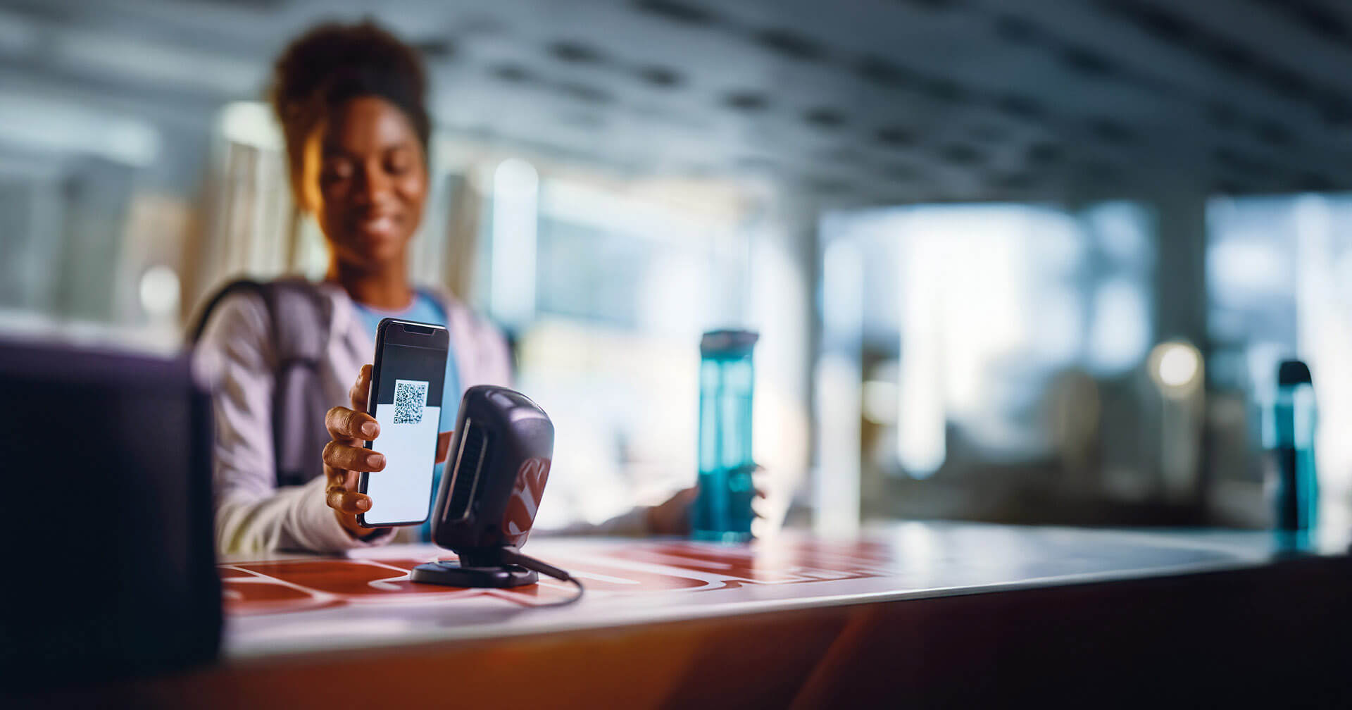 Eine junge Frau checkt per Smartphone in einem Fitnessstudio ein, indem sie einen QR-Code vor einen Scanner am Tresen hält. Der Hintergrund ist hell und modern, aber unscharf gehalten.