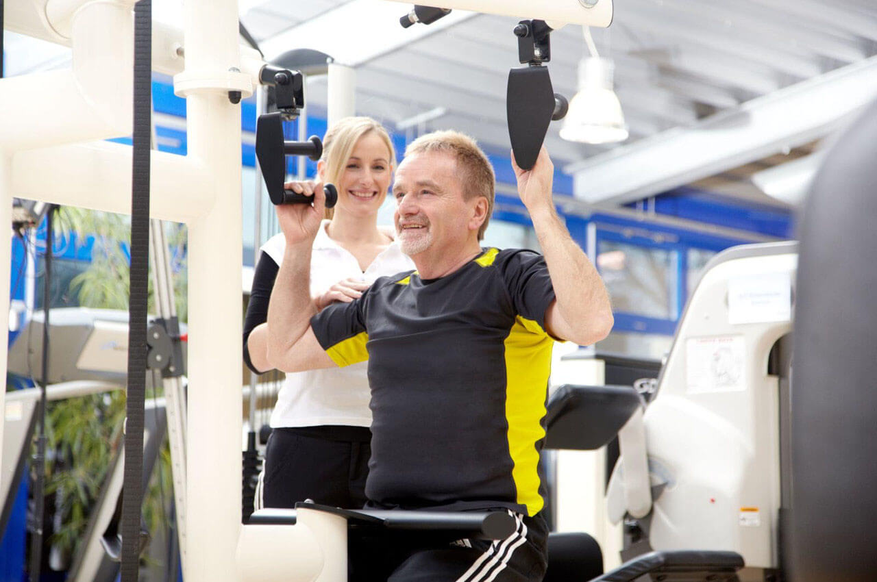 Älterer Mann trainiert an einem Fitnessgerät und wird dabei von einer Trainerin unterstützt, beide lächeln.