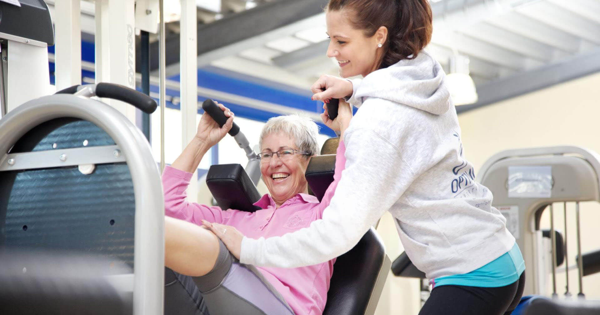 Ältere Frau trainiert lachend an einem Fitnessgerät, unterstützt von einer jungen Trainerin im Fitnessstudio.