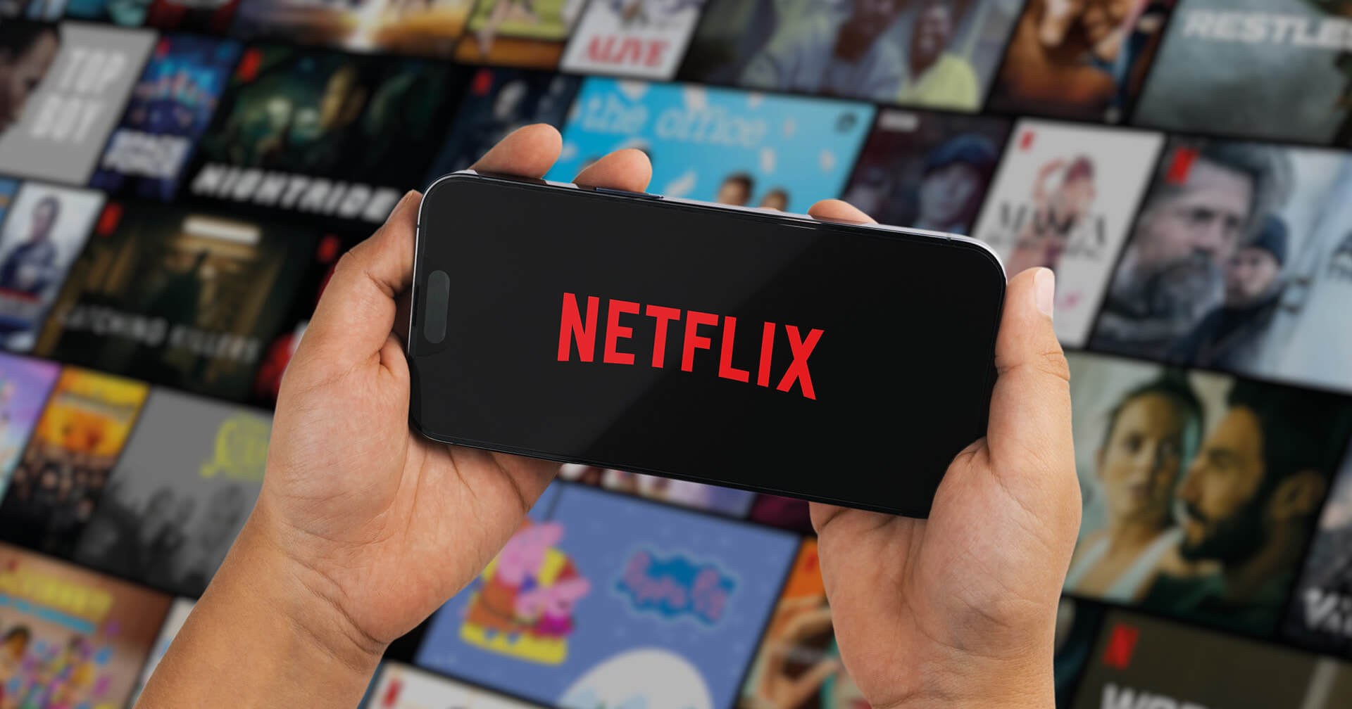 Nahaufnahme von zwei Händen, die ein Smartphone waagerecht halten. Auf dem schwarzen Bildschirm leuchtet das rote Netflix-Logo. Der Hintergrund zeigt eine unscharfe Galerie mit zahlreichen Film- und Serien-Vorschaubildern.