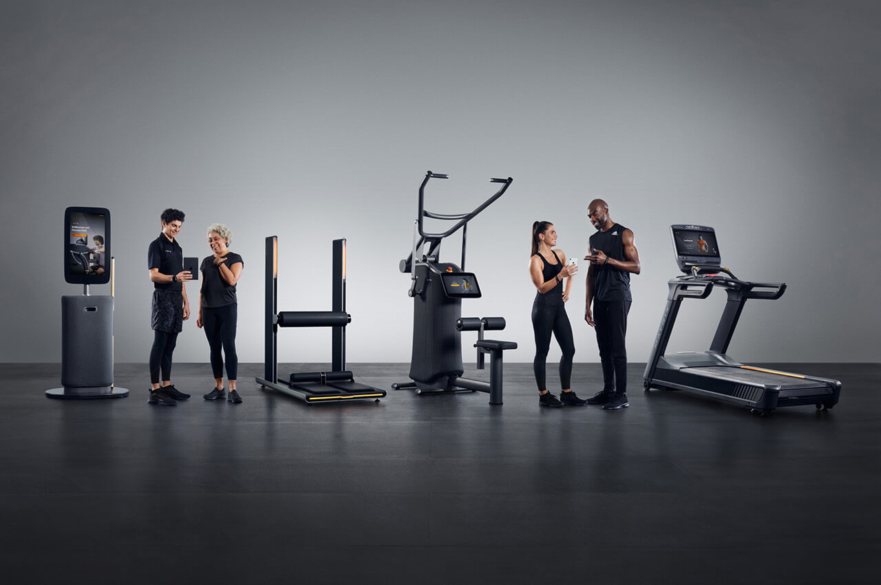  Hier ist ein Alt-Text für das Bild:  Eine Gruppe von vier sportlich gekleideten Personen interagiert in einem Fitnessstudio mit verschiedenen Geräten der EGYM Produktpalette.