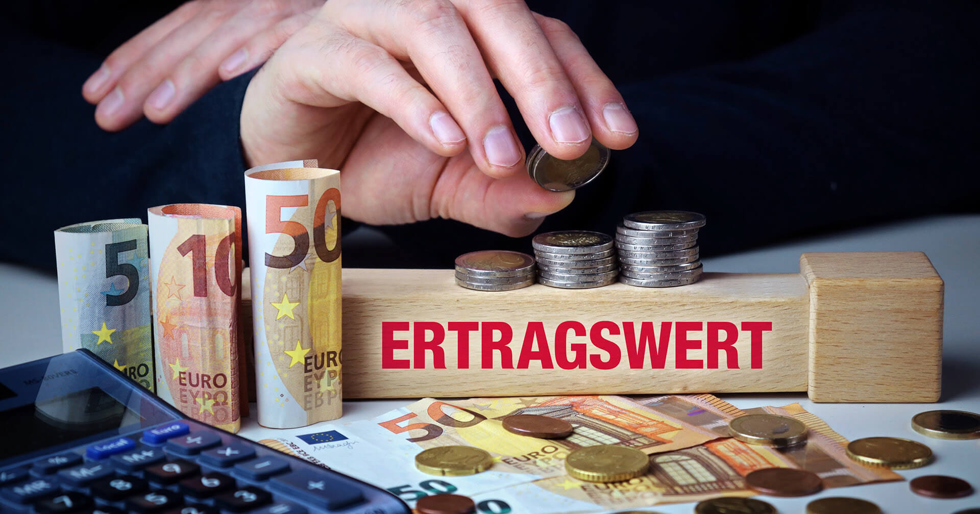 Eine Hand in dunkler Kleidung legt eine Münze auf einen Stapel Münzen, die auf einem Holzbalken vor Euro-Banknoten und einem Taschenrechner liegen. Auf dem Holzbalken steht in großen, roten Buchstaben das Wort "ERTRAGSWERT".
