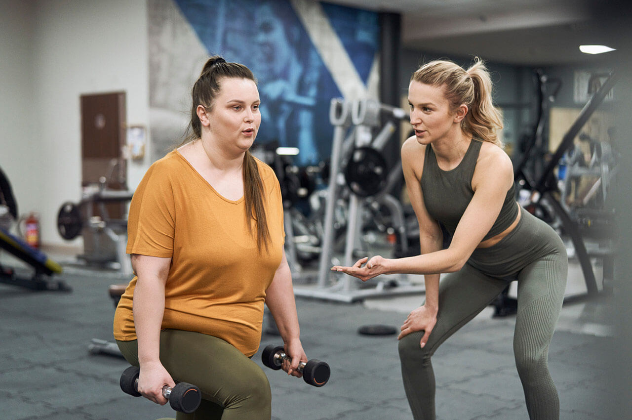  Eine Trainerin in olivgr&uuml;ner Sportkleidung unterst&uuml;tzt eine korpulente Frau im senfgelben T-Shirt bei Ausfallschritten mit Kurzhanteln. Beide befinden sich in einem Fitnessstudio mit Kraftger&auml;ten im Hintergrund. Die Trainerin erkl&auml;rt die &Uuml;bung empathisch.