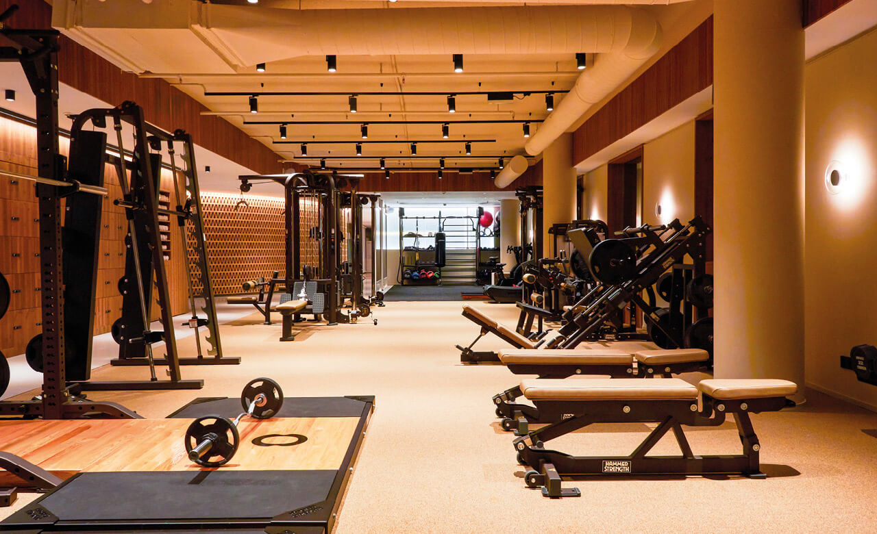 Exklusiv gestalteter Kraftraum mit Hantelbänken, Gewichten und modernen Fitnessgeräten in warm beleuchtetem Ambiente.