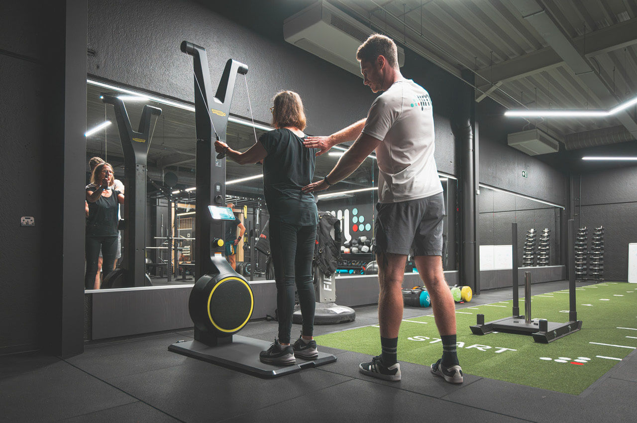 Healthness in der Fitnessbranche: Das ganzheitliche Konzept der Kette Jims - 1 Ein männlicher Coach unterstützt eine ältere Frau an einem modernen Krafttrainingsgerät mit digitalem Display. Er legt ihr korrigierend die Hand auf den Rücken, um eine gesunde Haltung sicherzustellen. Das Studio ist dunkel gestaltet mit hellem Holzboden.