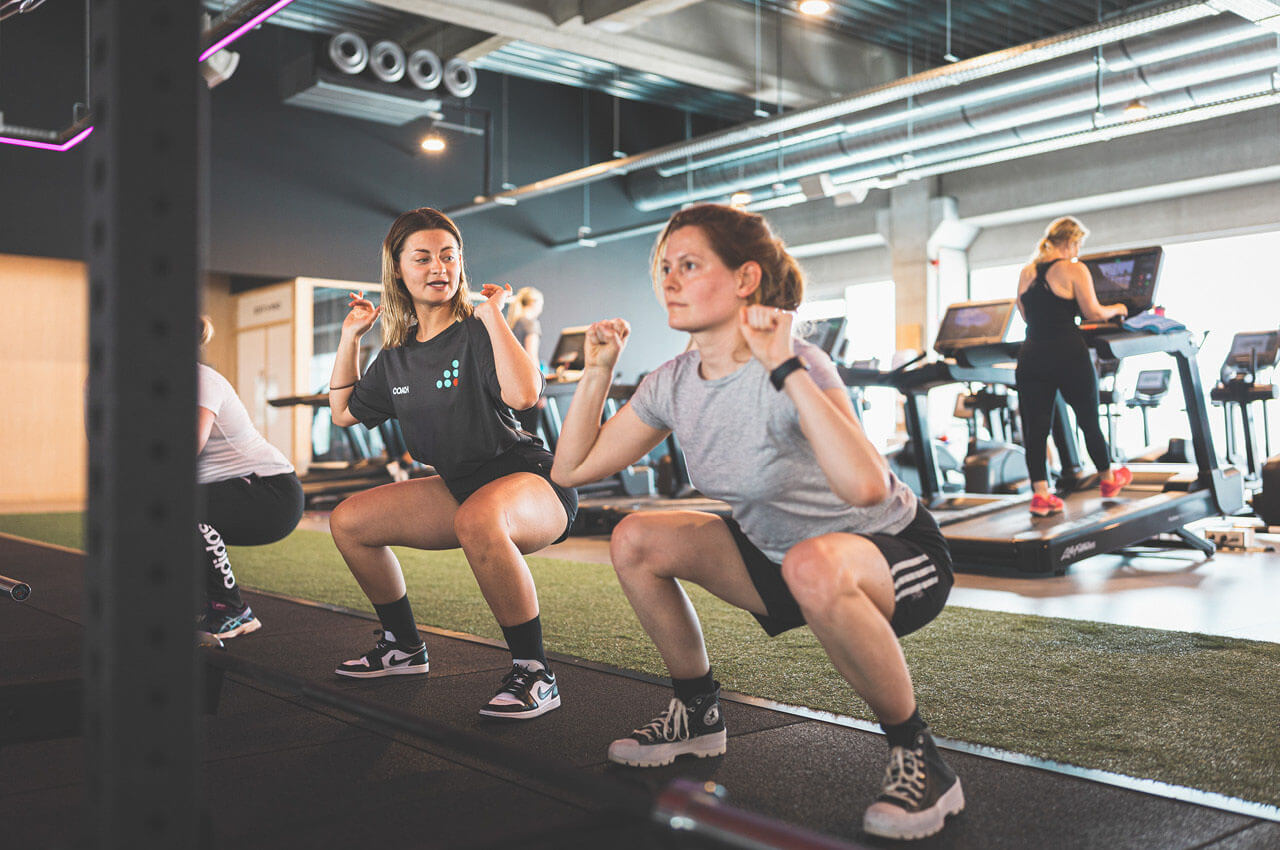Healthness in der Fitnessbranche: Das ganzheitliche Konzept der Kette Jims - 2 Ein Trainer von Jims im schwarzen T-Shirt mit Logo korrigiert eine junge Frau bei einer tiefen Kniebeuge. Beide befinden sich in einem modern ausgestatteten Fitnessstudio mit Kunstrasen-Laufbahn im Hintergrund.