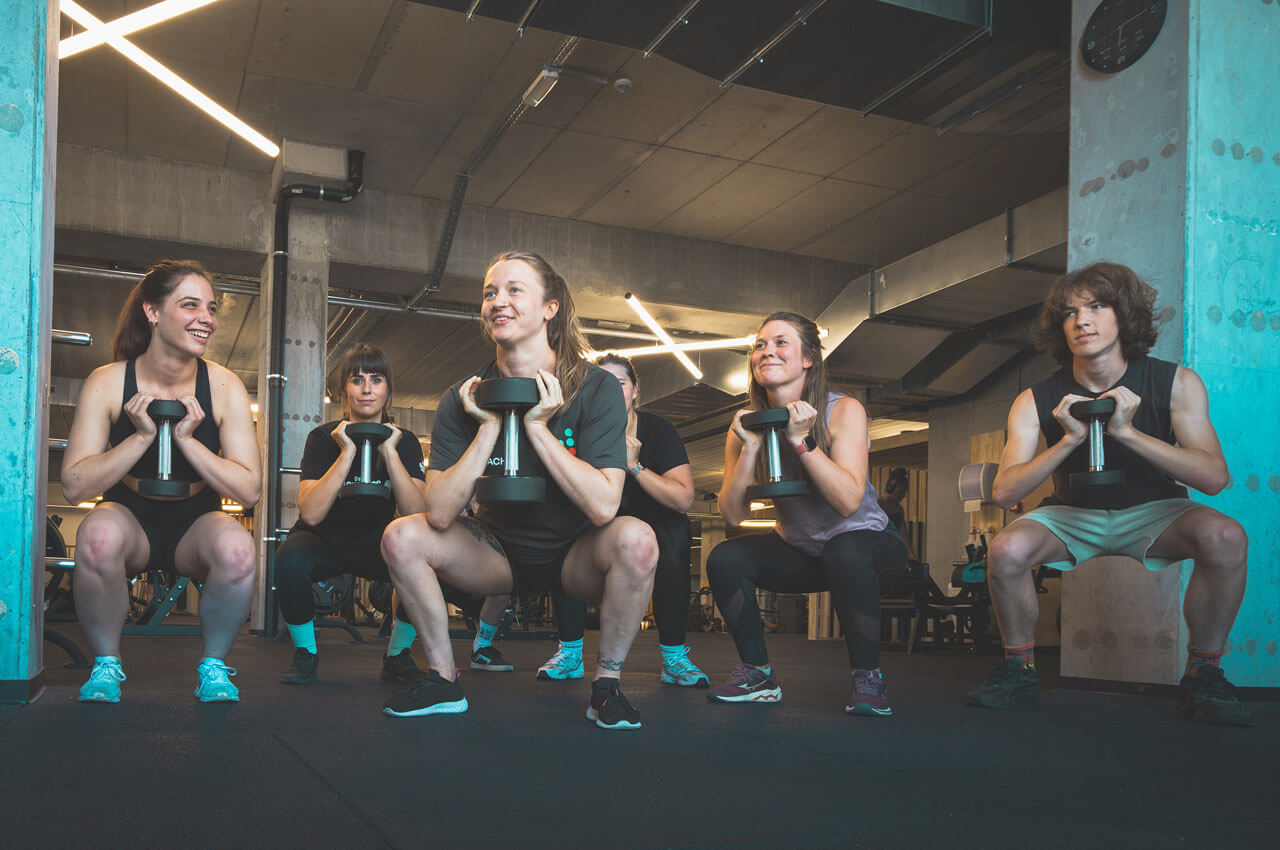 Healthness in der Fitnessbranche: Das ganzheitliche Konzept der Kette Jims - 3 Eine Gruppe von fünf jungen Erwachsenen führt synchron Kniebeugen mit Kurzhanteln aus. Sie lächeln sich gegenseitig an. Der Raum ist im industriellen Stil mit Betonpfeilern und moderner Lichtinstallation gestaltet.