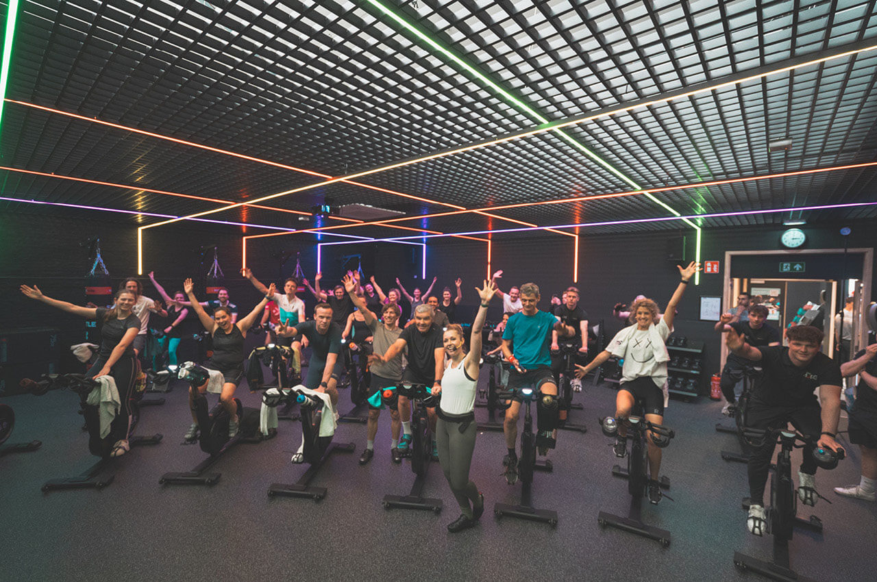 Healthness in der Fitnessbranche: Das ganzheitliche Konzept der Kette Jims - 4 Eine große Gruppe von Menschen trainiert begeistert in einem Indoor-Cycling-Kurs. Viele Teilnehmende haben die Arme jubelnd in die Luft gestreckt. Die Decke ist mit einem Raster aus bunten LED-Lichtstreifen in Orange, Grün und Violett gestaltet.