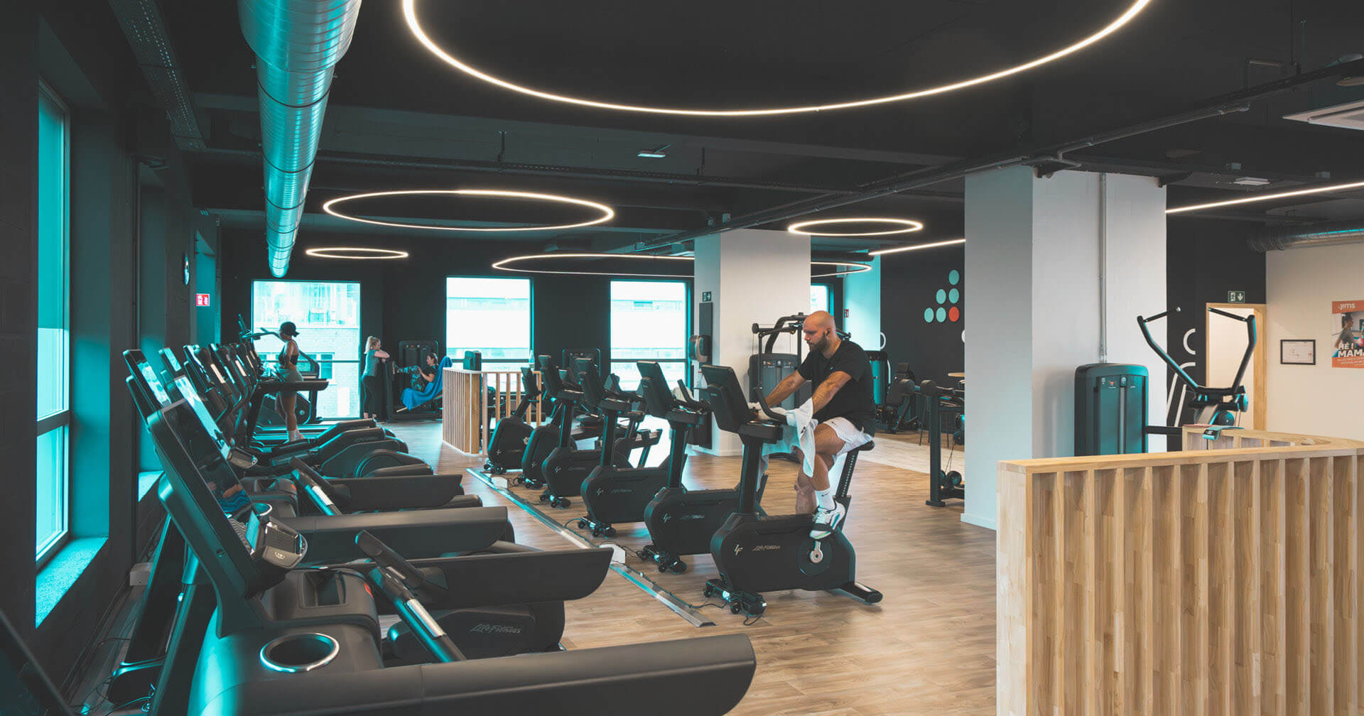 Weitwinkelaufnahme eines modernen, dunkel gestalteten Fitnessstudios. Ein Mann trainiert im Vordergrund auf einem Ergometer. Im Hintergrund sind Reihen von Laufbändern, weitere Kardiogeräte und Kraftstationen zu sehen. Der Raum ist durch auffällige, kreisrunde LED-Leuchten an der Decke und türkisfarbene Lichtakzente an den Fenstern beleuchtet. Holzverkleidungen setzen natürliche Akzente.