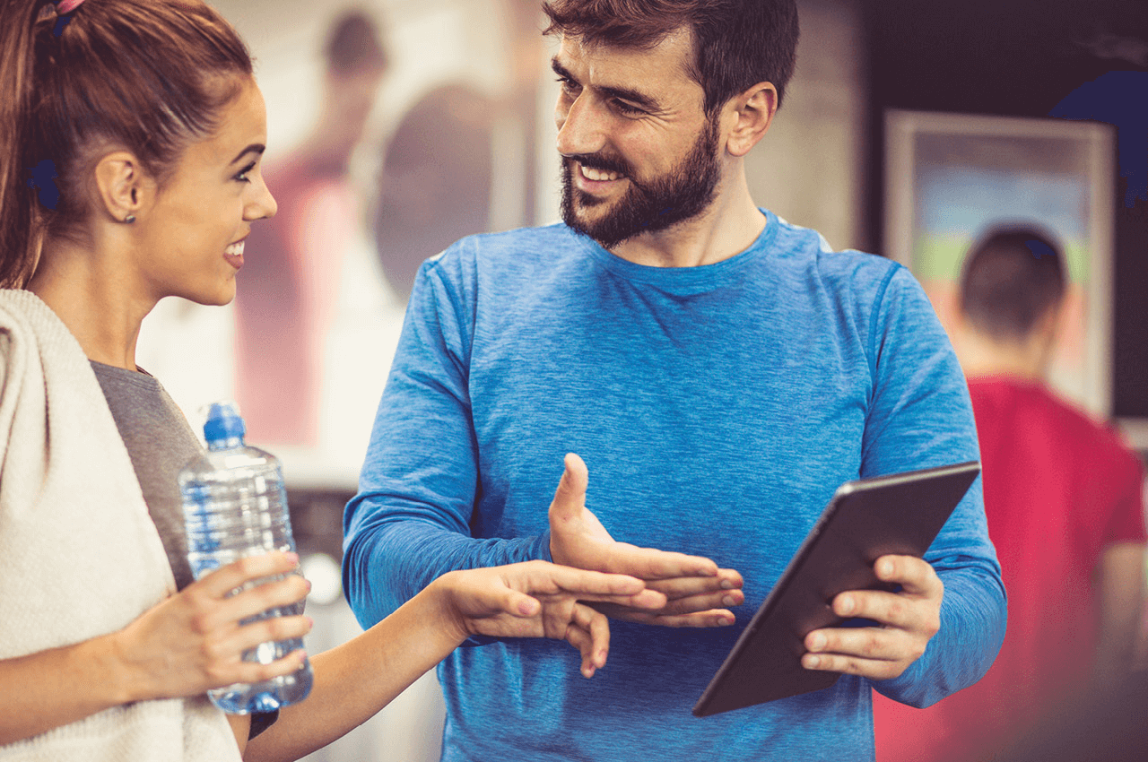Ein Trainer hat ein Tablet in der Hand und spricht mit einer jüngeren Trainierenden in einem Fitnessstudio