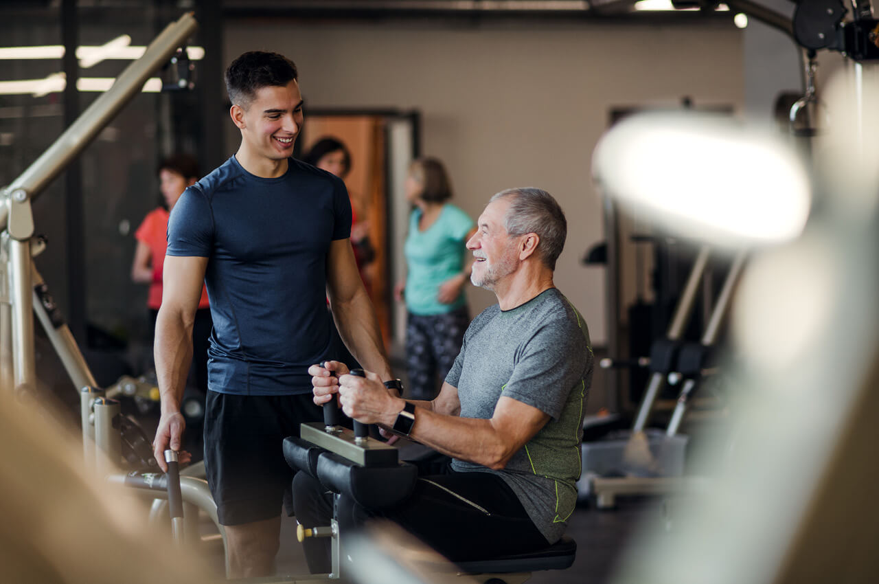 Junge Mitarbeiter freuen sich, wenn sie erleben, dass ihre Energie Wirkung entfaltet. Von dieser Energie profitieren auch Kunden Junger Trainer steht vor einem älteren Herrn, der im Fitnesscenter gerade auf einer Maschine trainiert