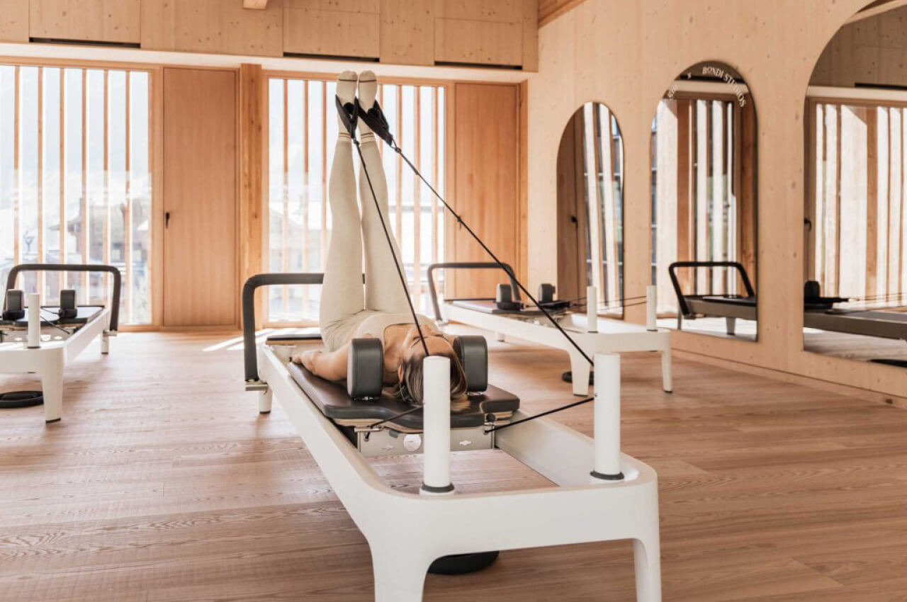 Eine Frau liegt auf einem Pilates-Reformer in einem hellen Holzstudio. Ihre Beine sind senkrecht nach oben gestreckt, die F&uuml;&szlig;e in den Schlaufen der Seilz&uuml;ge. Sie konzentriert sich auf die &Uuml;bung, w&auml;hrend im Hintergrund weitere Reformer und Spiegel zu sehen sind.