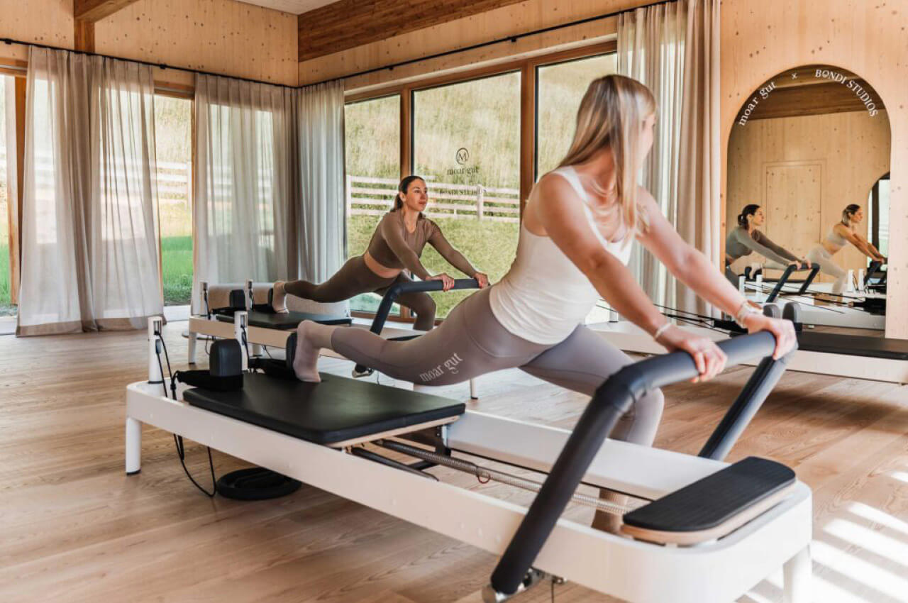 Zwei Frauen trainieren synchron auf Pilates-Reformern in einem hellen Studio mit Blick ins Gr&uuml;ne. Sie f&uuml;hren einen kraftvollen Ausfallschritt aus.