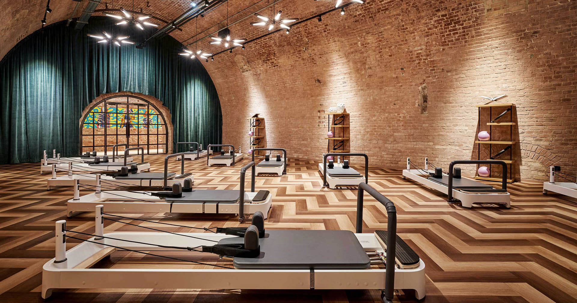 Innenansicht eines großen, stilvollen Pilates-Lofts. Der Raum hat gewölbte Ziegelwände, einen Fischgrät-Parkettboden und moderne Kronleuchter. Mehrere weiße Pilates-Reformer stehen in Reihen bereit. Am Ende des Raumes befindet sich ein großes Bogenfenster mit einem grünen Vorhang.