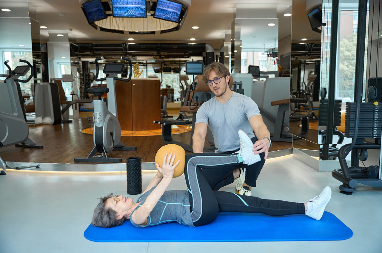 Trainer unterstützt eine ältere Frau bei einer Übung auf der Matte mit Medizinball in einem modernen Fitnessstudio.